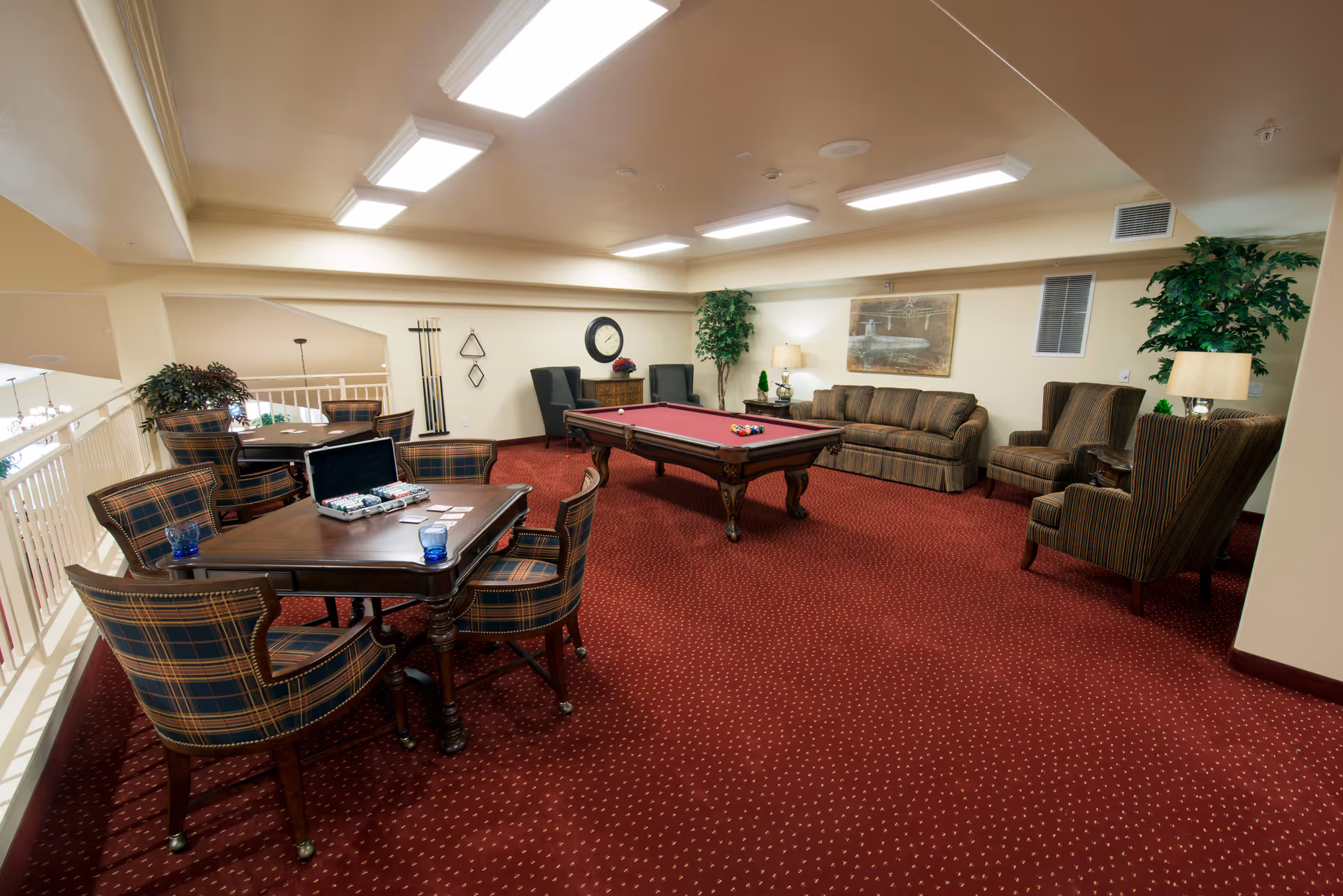 A spacious recreational room with a red carpet featuring small yellow dots. The room includes a pool table with balls set up, a seating area with a striped sofa and several armchairs, and a card table with four plaid upholstered chairs. The card table has poker chips, cards, and two blue glasses on it. The walls are light-colored with a clock, a painting, and two large potted plants. The ceiling has multiple rectangular fluorescent lights.