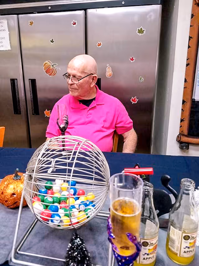 An elderly man wearing glasses and a bright pink polo shirt sits at a table with a bingo cage filled with colorful balls in front of him. The background shows a metallic refrigerator decorated with autumn-themed stickers such as pumpkins and leaves. On the table, there are several bottles, a glass of beer, and Halloween decorations including a glittery pumpkin and a black cat figure.