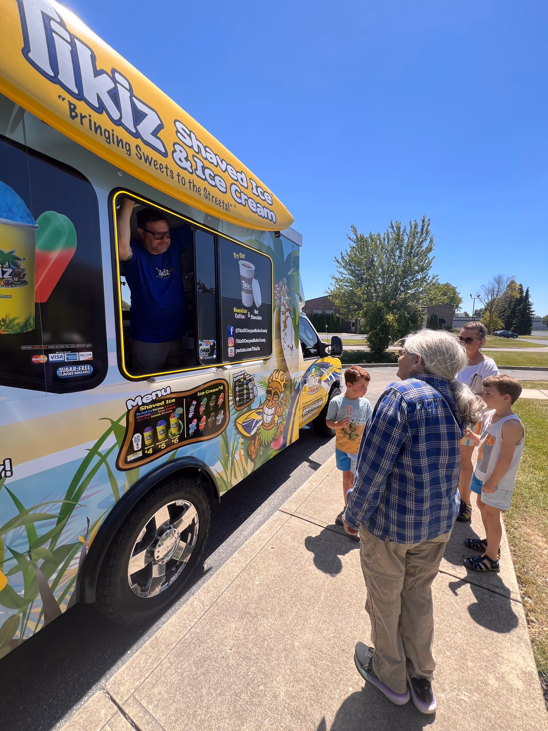 A colorful shaved-ice truck serving customers on a sunny sidewalk as several people wait.