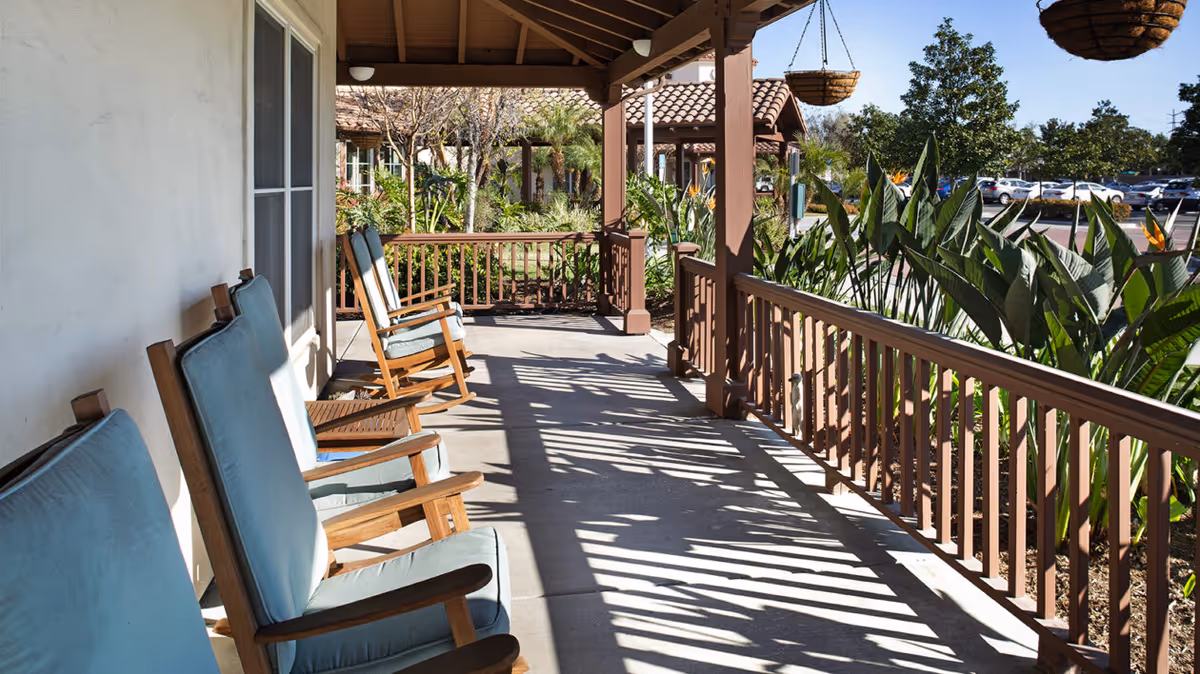Covered porch with cushioned wooden rocking chairs overlooking landscaped plants and a parking lot.
