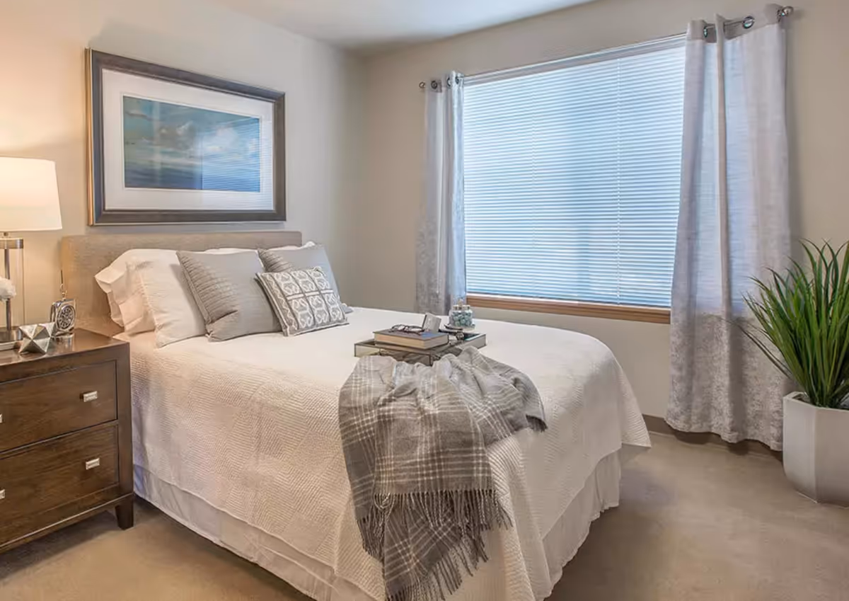 A neatly made bed with white bedding and multiple pillows in a bedroom. There is a wooden nightstand with a lamp and decorative items on the left side of the bed. A large window with closed blinds and light-colored curtains is on the right wall. A framed picture hangs above the bed, and a potted plant is placed near the window on the floor.
