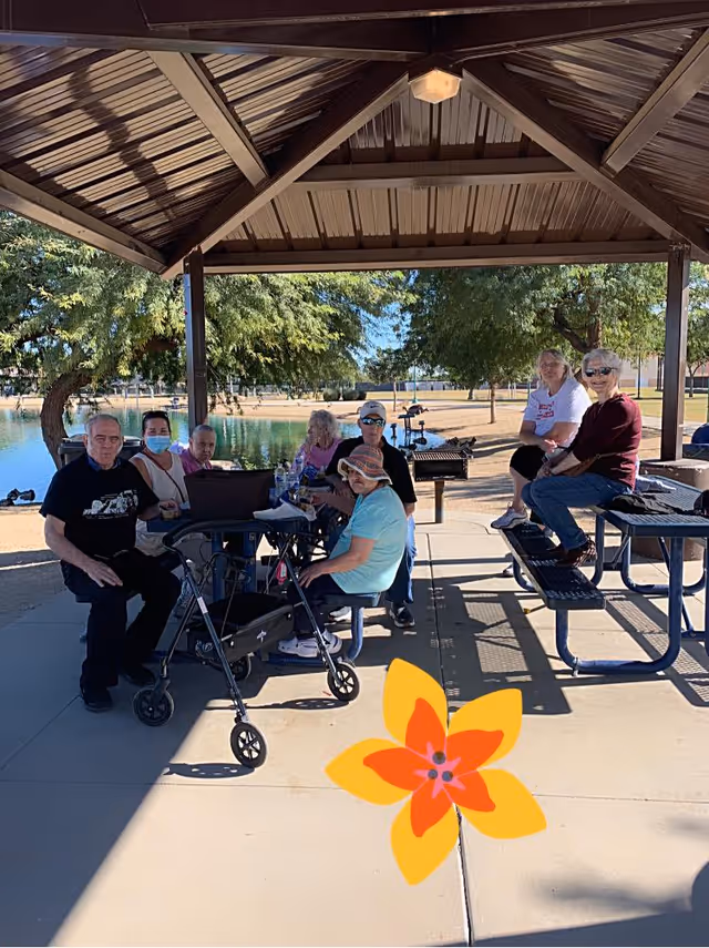A group of elderly people sitting and socializing under a shaded pavilion near a pond in a park. Some are seated at picnic tables while one person uses a walker. Trees and a clear blue sky are visible in the background.