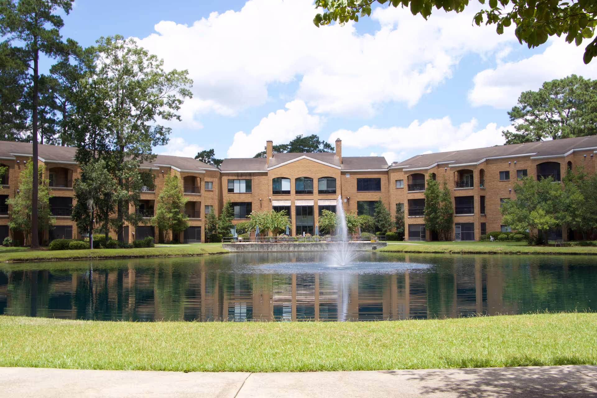 A large three-story brick building with multiple windows and balconies, situated behind a pond with a central water fountain. The building is surrounded by green trees and grass under a partly cloudy blue sky.