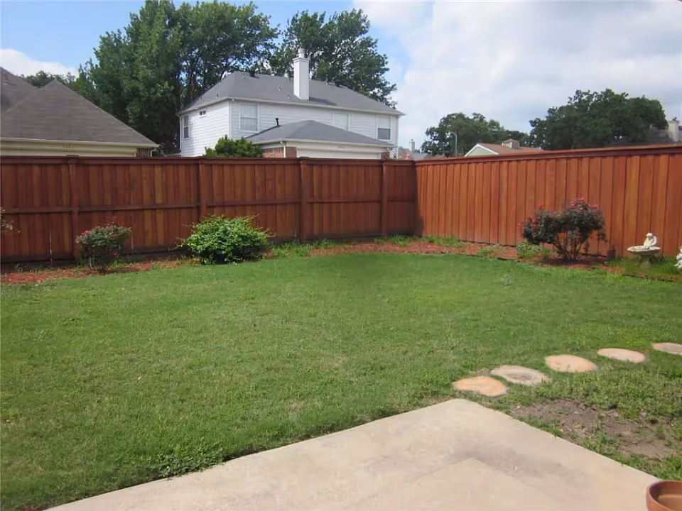 A backyard with a green lawn, a wooden fence surrounding the area, some small bushes and plants along the fence, and a stone pathway leading from a concrete patio. In the background, there are neighboring houses and trees under a partly cloudy sky.