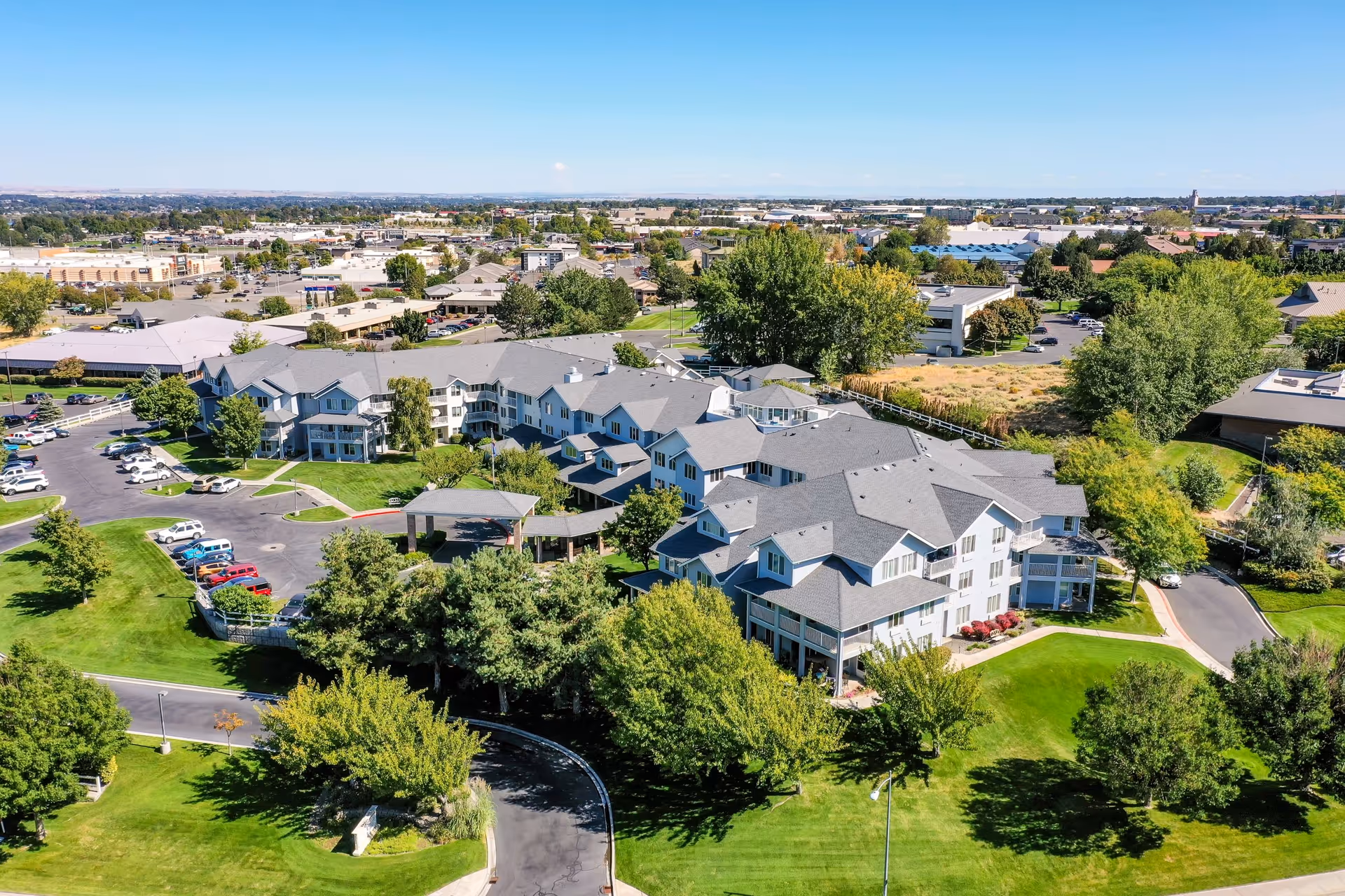 Aerial view of a senior living facility surrounded by green lawns, trees, and parking areas. The building has multiple wings with gray roofs and white exterior walls. The facility is situated in a suburban area with other buildings and roads visible in the background under a clear blue sky.