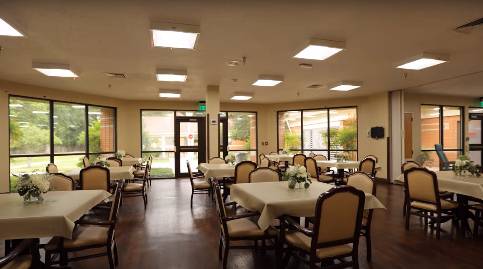 A spacious dining room with multiple tables covered in beige tablecloths, each adorned with a small floral centerpiece. The room has large windows allowing natural light to enter, and several chairs are arranged around the tables. The floor is wooden, and the ceiling has multiple recessed lights.