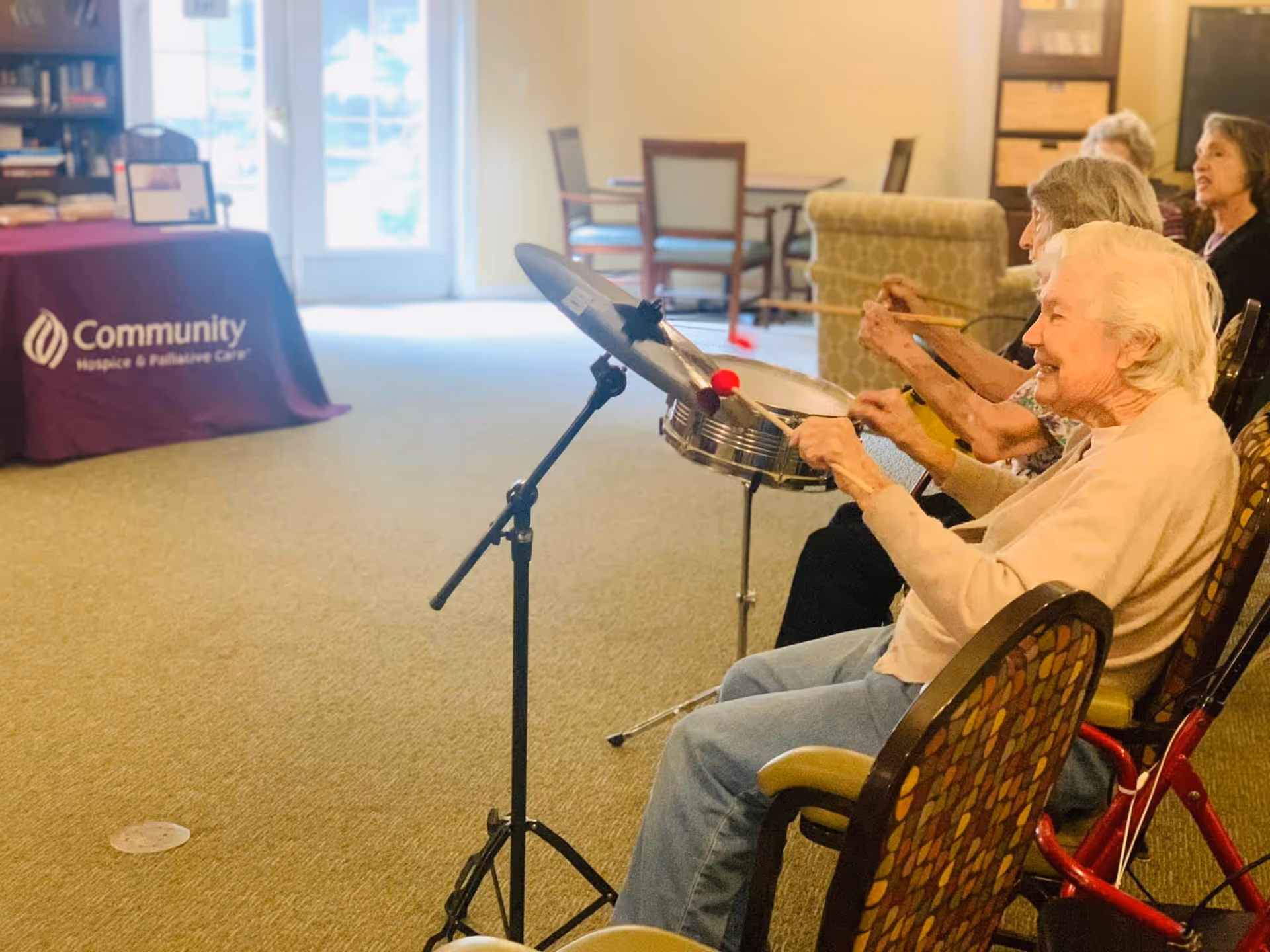A group of elderly women seated in chairs inside a senior living facility, playing percussion instruments with drumsticks. The room has carpeted flooring, a table with a purple cloth that reads 'Community Hospice & Palliative Care,' and some furniture in the background.