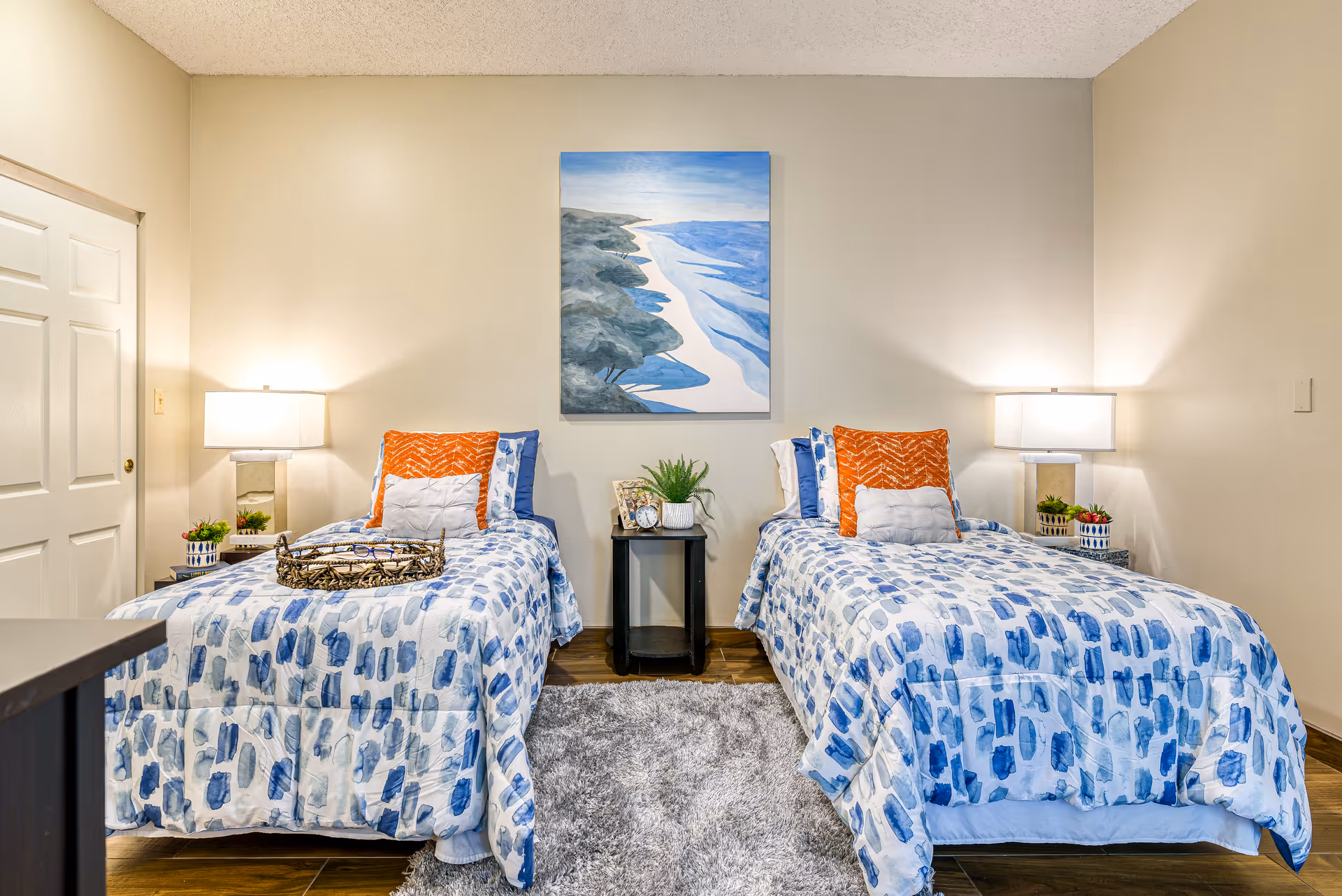 Two twin beds with blue-patterned bedding and orange accent pillows in a staged bedroom with bedside lamps and a coastal painting.