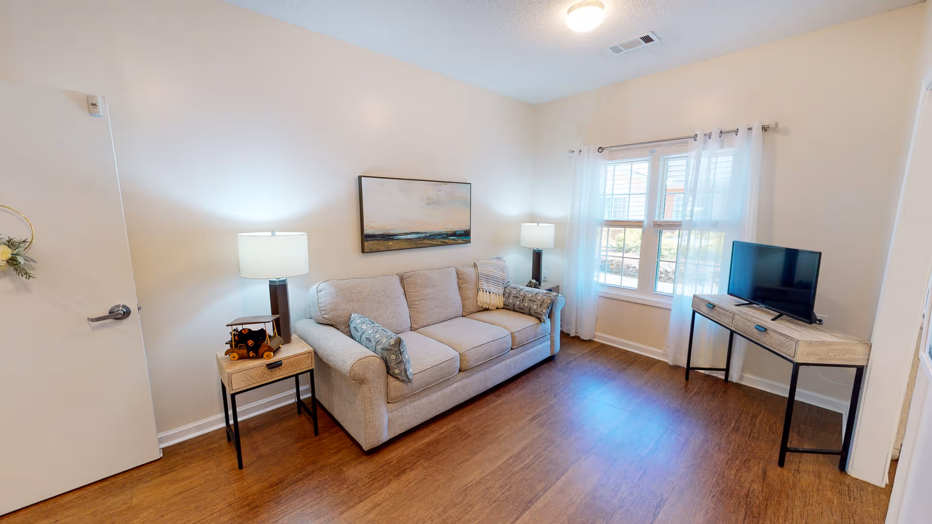 Small furnished living room with a beige sofa, side tables and lamps, a TV on a console, and a window with sheer curtains.