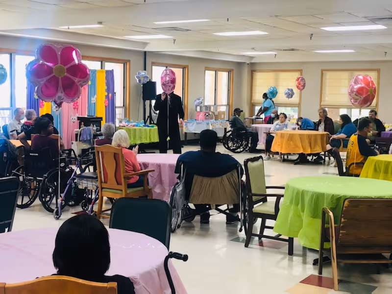 Residents, some in wheelchairs, sit at round tables with colorful tablecloths and balloons in a decorated dining/activity room while a person speaks at the front.