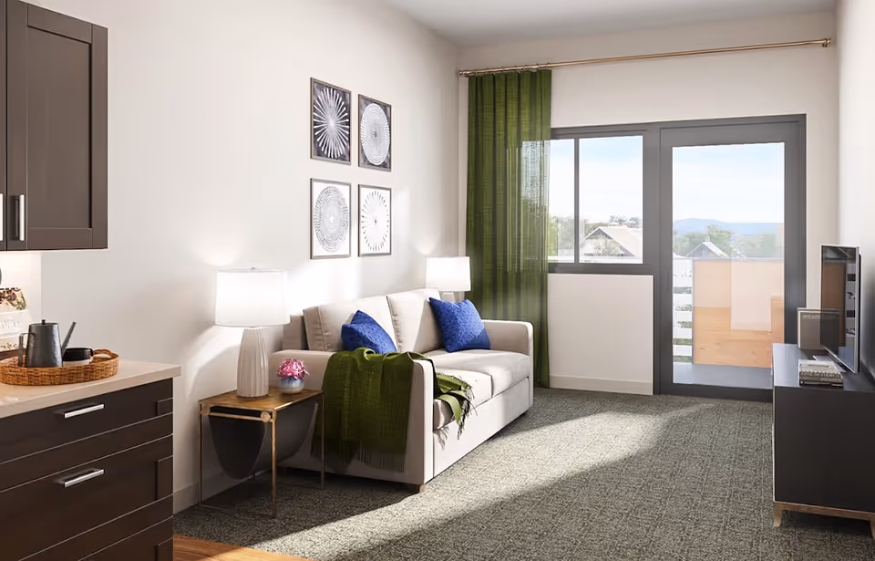 A bright living room with a beige sofa adorned with two blue pillows and a green throw blanket. Next to the sofa is a wooden side table with a white lamp and a small flower vase. The room has a large window and a glass door leading to a balcony with a view of rooftops and distant hills. On the left side, there are dark brown kitchen cabinets and a countertop with a tray holding a teapot and cups. A TV is placed on a dark TV stand on the right side of the room. The walls are decorated with four framed abstract art pieces, and green curtains hang by the window and door.