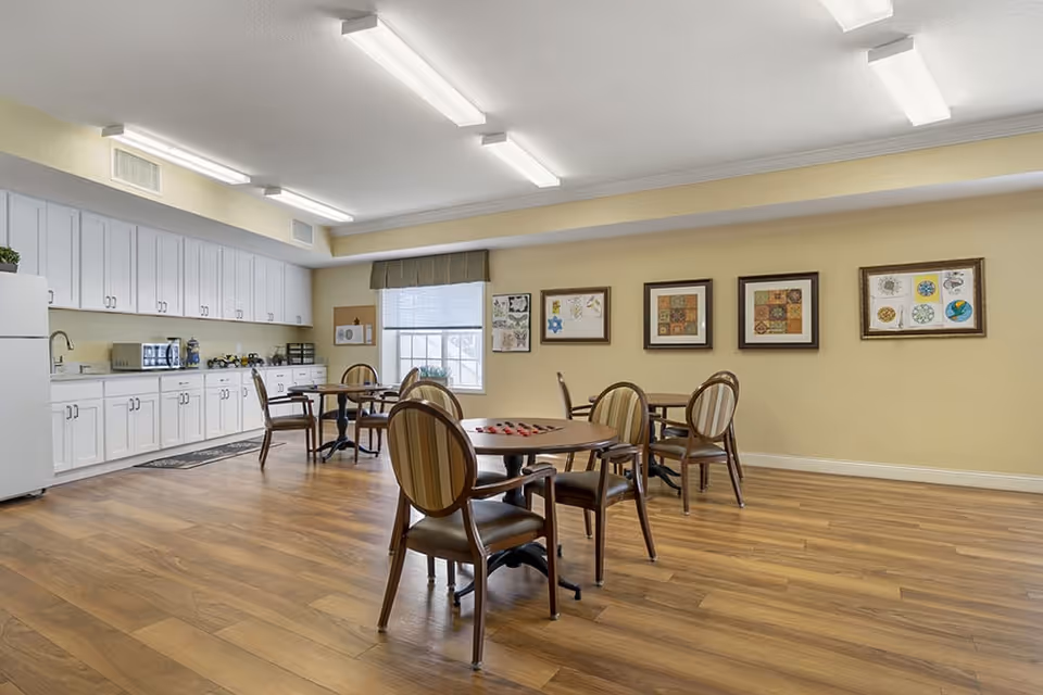 Bright communal dining/activity room with round tables and chairs, a kitchenette with white cabinets and appliances, and framed artwork on the wall.