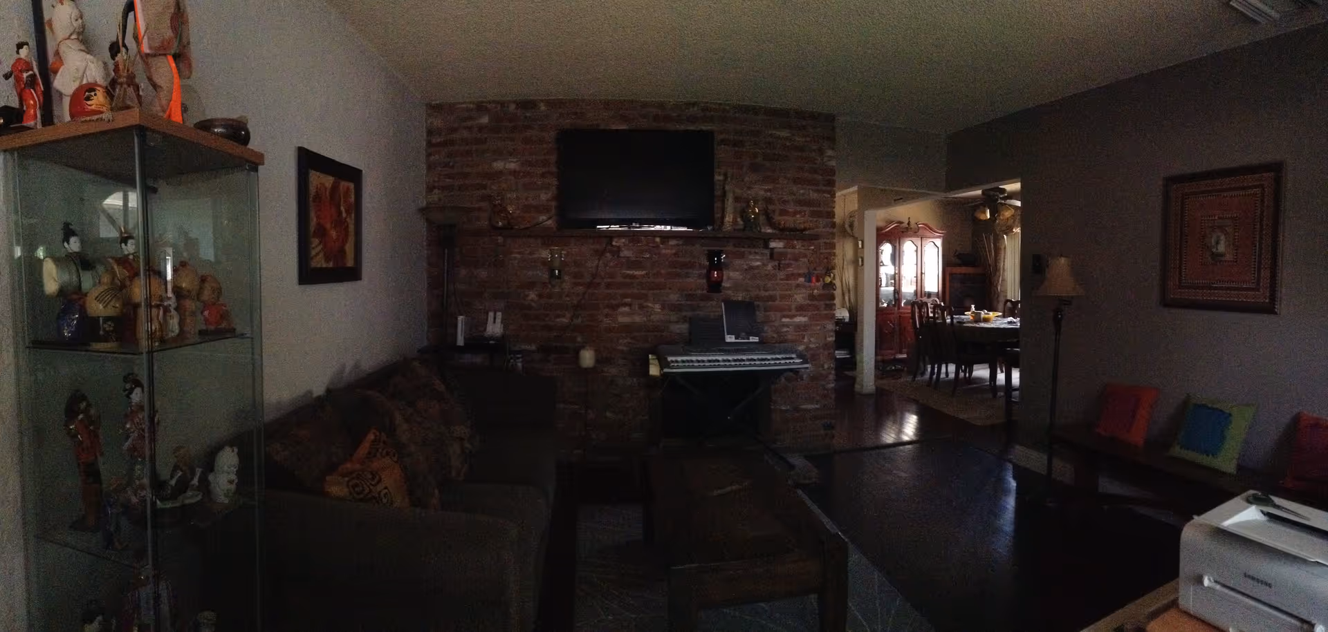 A cozy living room with a dark sofa adorned with patterned cushions, a wooden coffee table, and a brick wall with a mounted flat-screen TV above a shelf holding decorative items. To the left, a glass display cabinet contains various figurines. The room opens into a dining area with a wooden dining table and chairs, a china cabinet, and a ceiling fan. There is a floor lamp and framed artwork on the walls.
