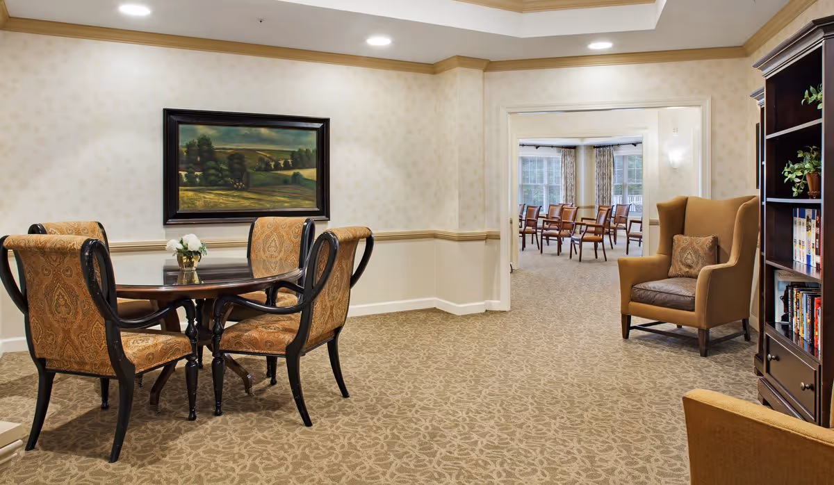 A cozy interior room with a round wooden table surrounded by four upholstered chairs with patterned fabric. A small vase with white flowers sits on the table. On the wall behind the table is a framed landscape painting. To the right, there is a comfortable wingback chair with a decorative pillow, and a dark wooden bookshelf filled with books and plants. Through an open doorway, another room is visible with several wooden chairs arranged in rows near large windows with curtains.