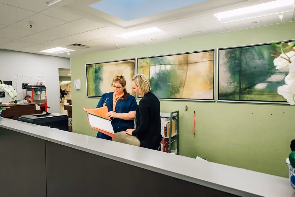 Two women standing behind a reception desk in a healthcare facility. One woman is wearing a blue uniform and holding an open binder, while the other woman, dressed in black, is looking at the binder. The background features a green wall with three abstract paintings and some office supplies.