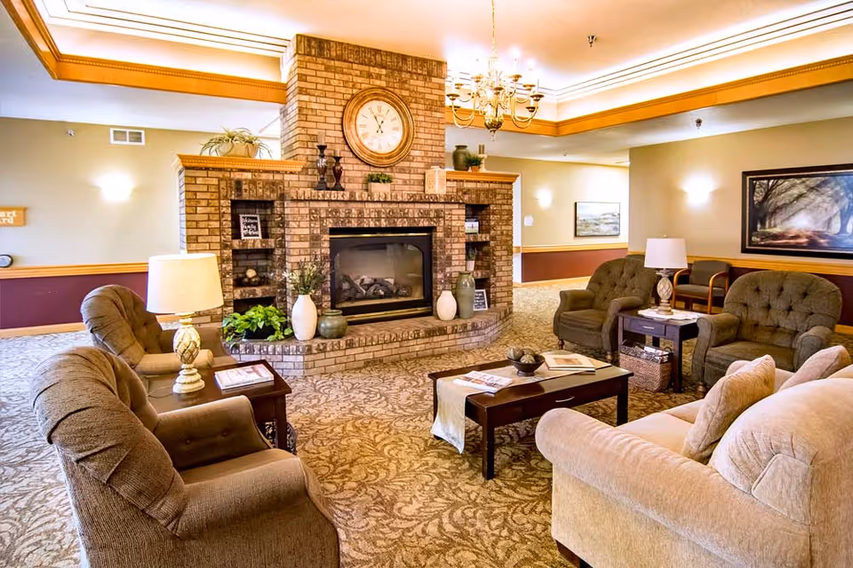 A cozy living room area featuring a brick fireplace with a large clock above it. The room is furnished with a beige sofa, two brown armchairs, a coffee table with books and decor, and side tables with lamps. The walls are painted in warm tones with framed artwork, and the carpet has a floral pattern. The ceiling has recessed lighting and a chandelier.