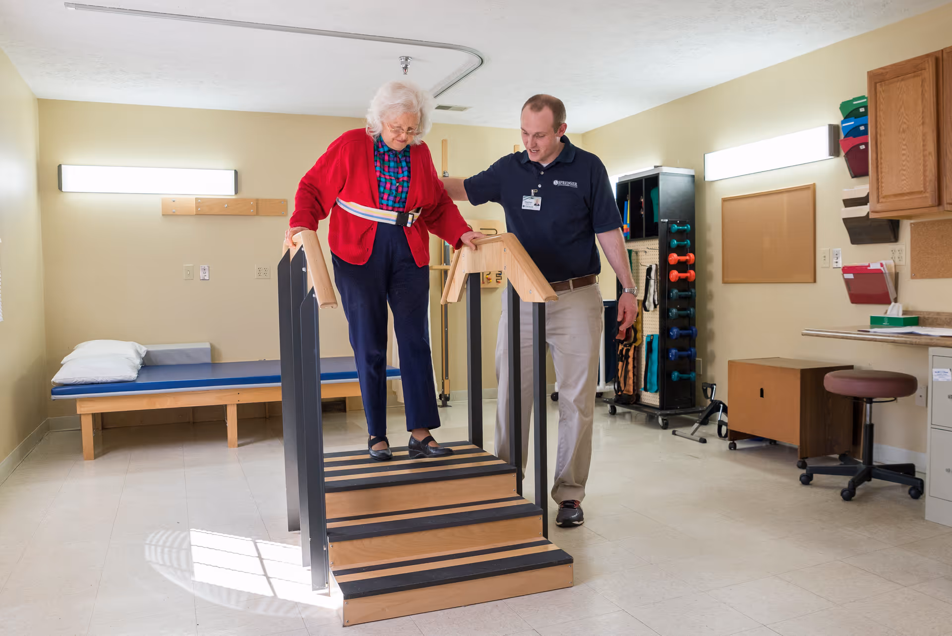 An elderly woman wearing a red cardigan and navy pants is walking down a small set of wooden stairs with handrails on both sides, assisted by a male healthcare professional in a navy shirt and khaki pants in a therapy room. The room has a padded bench with a pillow, exercise equipment including dumbbells, and office furniture.