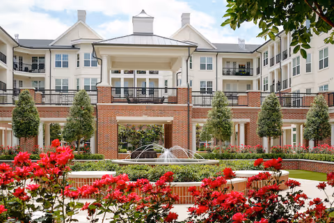 A landscaped courtyard with a central water fountain surrounded by red flowering bushes and greenery, in front of a multi-story senior living facility building with balconies and a covered seating area.