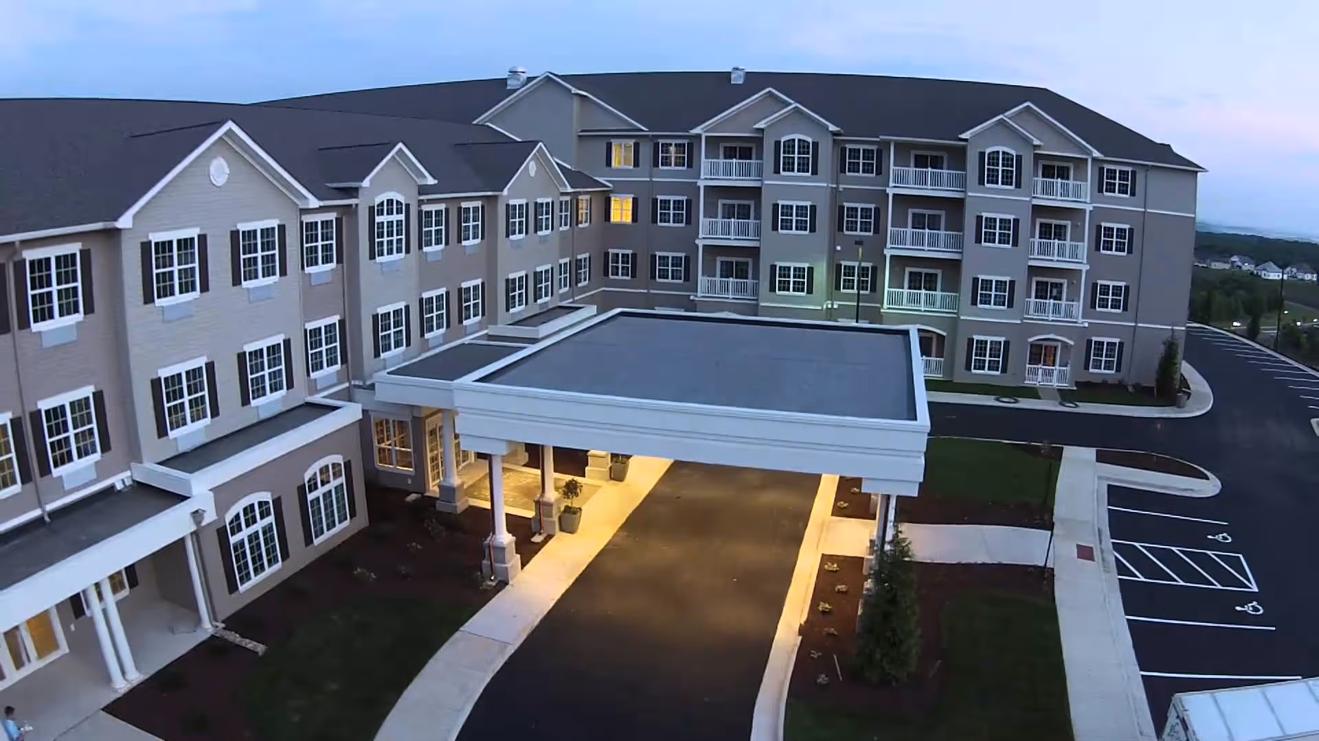 Exterior view of a multi-story senior living facility building at dusk with a covered entrance driveway, multiple windows, balconies, and a parking area with marked spaces including handicapped spots.