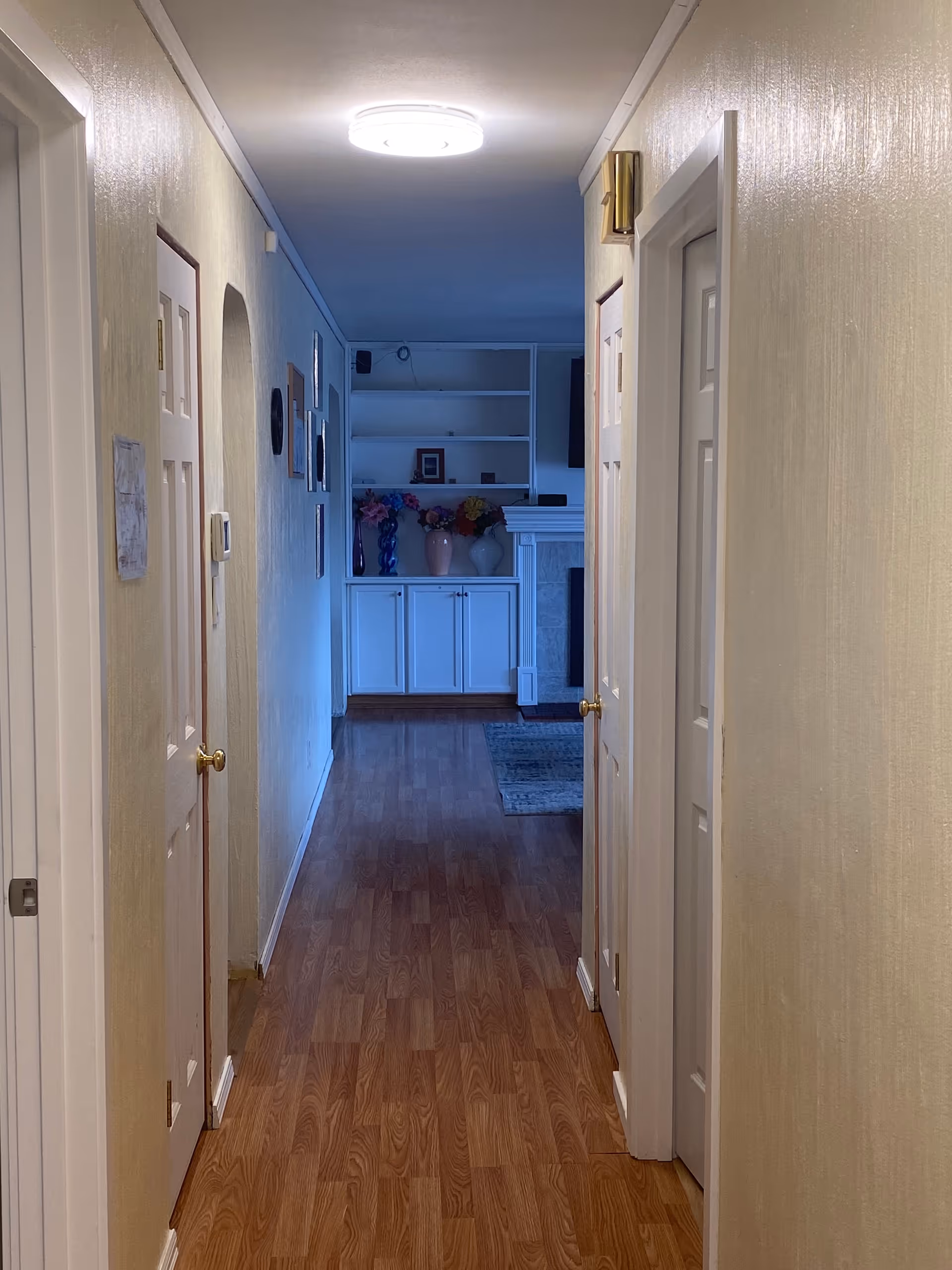 A hallway with wooden flooring and cream-colored walls leading to a living area with white cabinets, decorative vases with flowers, and a fireplace. Several closed doors line the hallway, and framed pictures hang on the left wall. A round ceiling light illuminates the hallway.