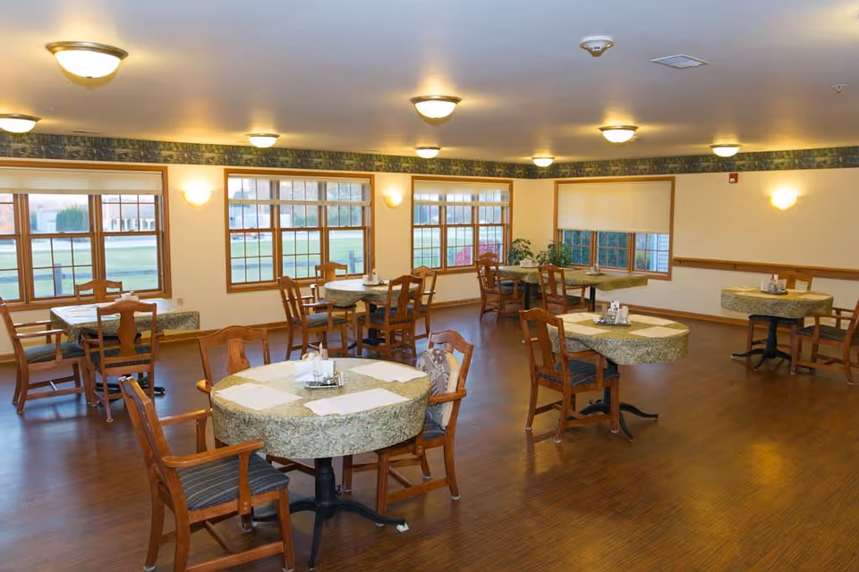 Bright dining room with round tables covered in patterned tablecloths, wooden chairs, large windows, and ceiling lights.