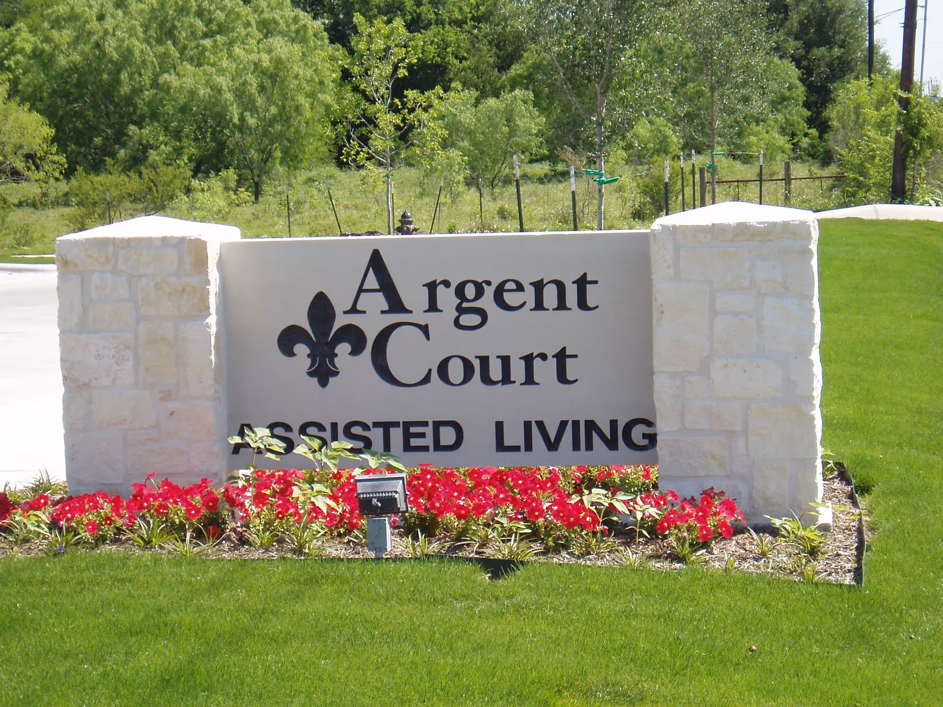 Stone sign for Argent Court Assisted Living surrounded by green grass and red flowers, with trees and a clear sky in the background.