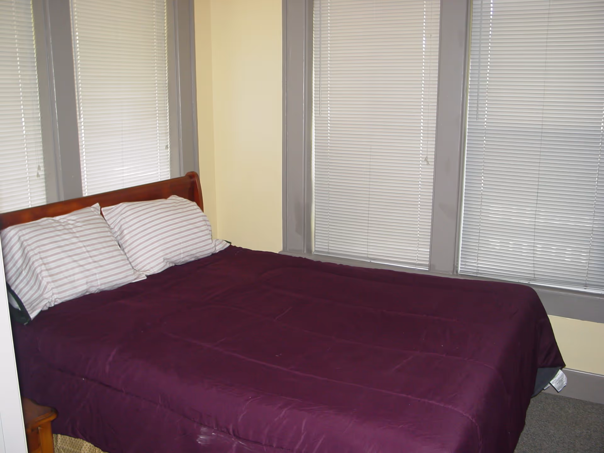 A bedroom with a wooden bed frame, a purple comforter, and two striped pillows. The room has three windows with closed white blinds and gray trim, and the walls are painted light yellow.