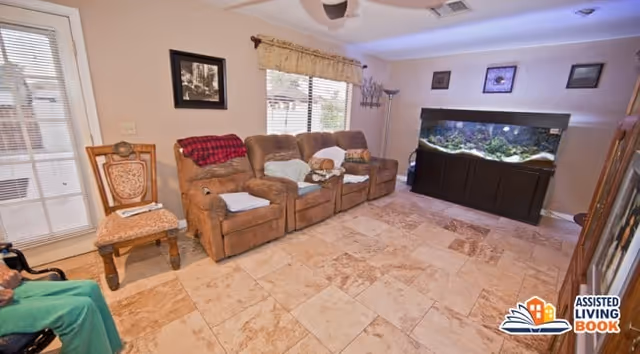 A cozy living room with three brown recliner chairs arranged in a row, each with a white cushion. A small dog is resting on one of the chairs. There is a wooden chair with a patterned cushion next to the recliners. The room has a large fish tank on a dark wooden stand against the wall, with framed pictures hanging above it. The floor is tiled with beige and brown stone tiles, and there is a window with blinds and a valance letting in natural light. A person wearing green pants is partially visible sitting on the left side of the image.