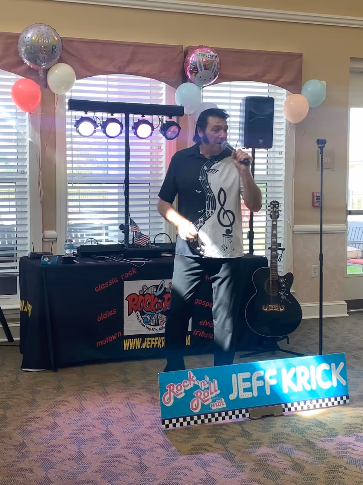 A man wearing a black and white shirt with musical notes is singing into a microphone in a decorated room with balloons and stage lights. Behind him is a table covered with a black cloth featuring the logo and website of Rock-n-Roll performer Jeff Krick, along with a guitar on a stand. The room has large windows with blinds and a carpeted floor.