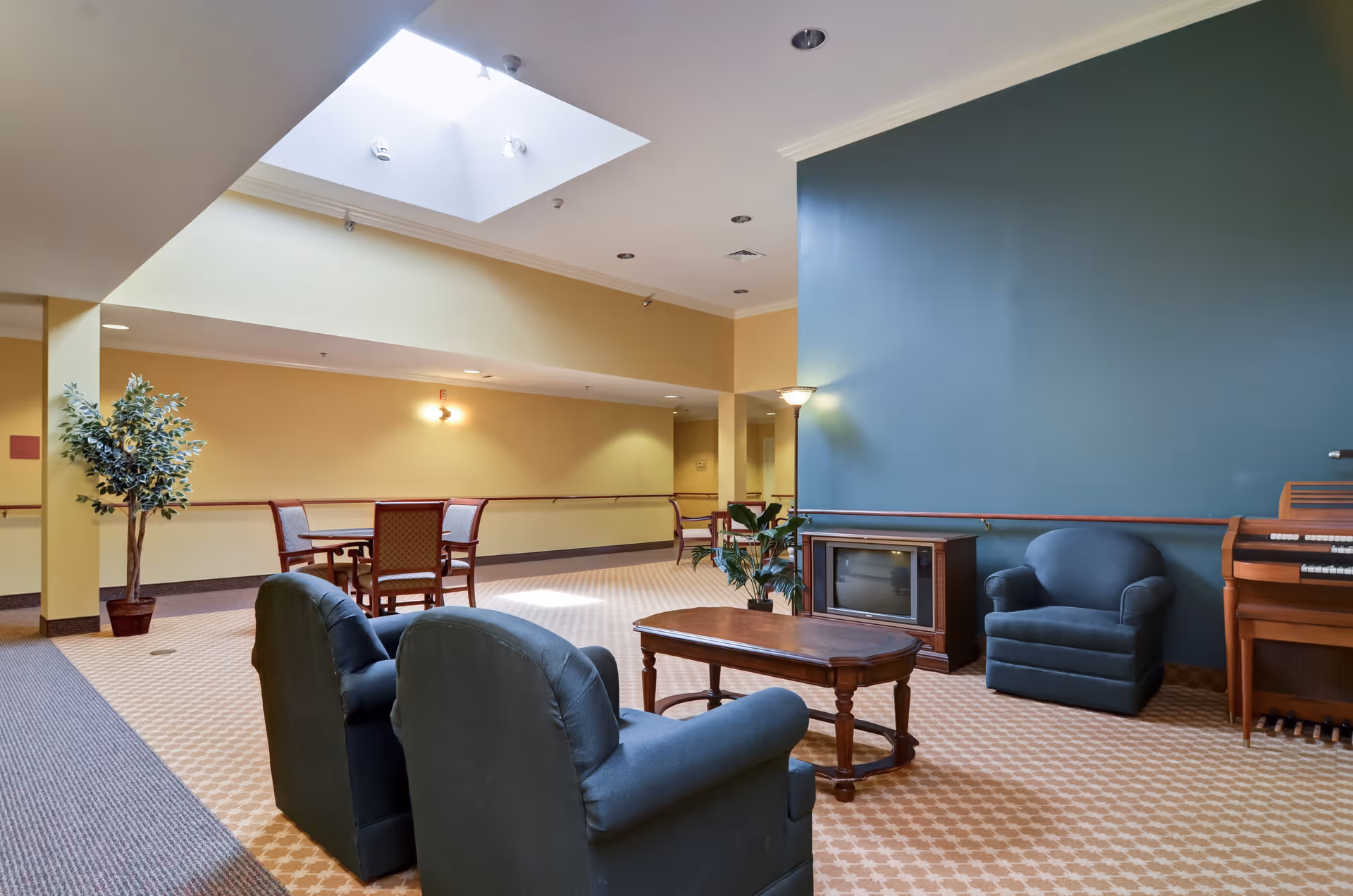 A common area in a senior living facility with two dark upholstered armchairs and a wooden coffee table in the foreground. There is a small vintage television on a wooden stand against a blue accent wall, next to a floor lamp and a wooden organ. In the background, there is a small table with four chairs and a potted plant near a yellow wall. The ceiling has a skylight allowing natural light to brighten the space.