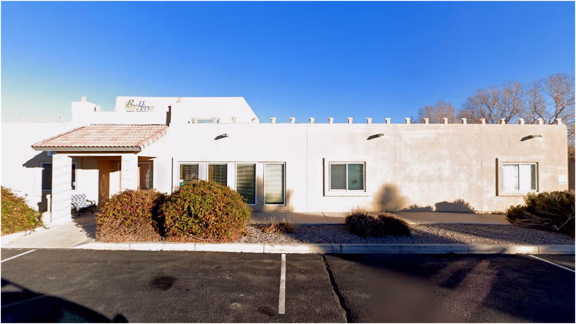Single-story stucco building front with an entrance, windows, low shrubs and a clear blue sky.