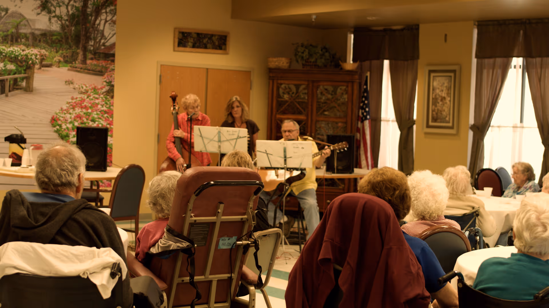 A group of elderly people seated in a common room, watching a small musical performance by three musicians playing guitar and double bass. The room has round tables, chairs, and large windows with curtains.