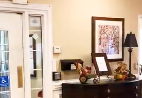 Interior view of a senior living facility entrance area showing a glass door with a handicap accessibility sign, a dark wooden console table with decorative items including a lamp, framed artwork on the wall, and small seasonal decorations.