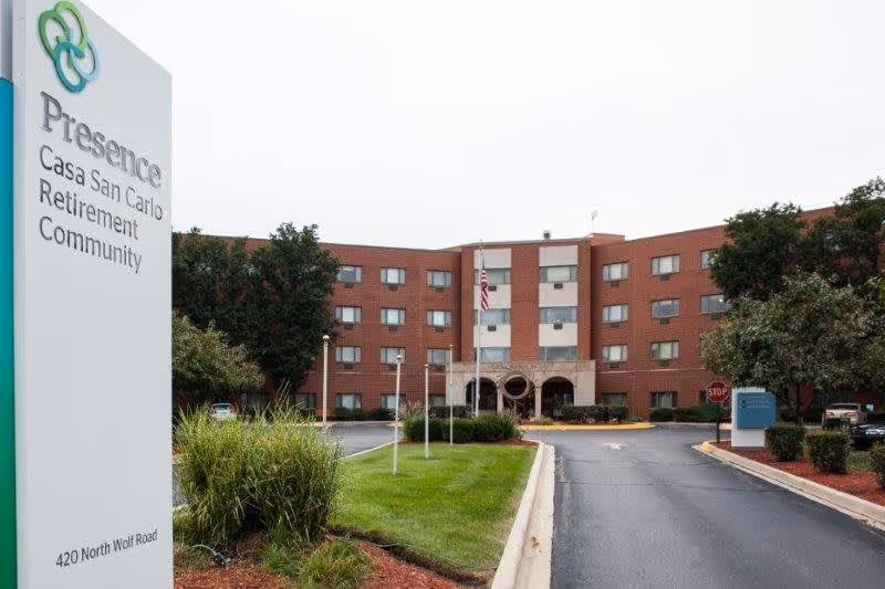 Exterior view of a multi-story brick retirement community building with an entrance driveway, green lawn, and trees. A tall sign in the foreground reads 'Presence Casa San Carlo Retirement Community' with the address 420 North Wolf Road. The sky is overcast.