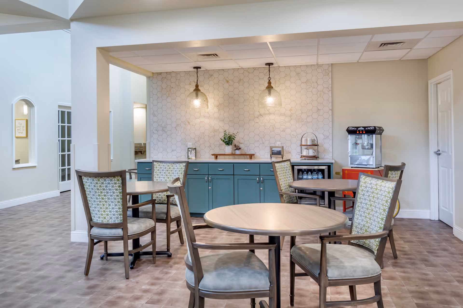 A bright and clean dining area with round wooden tables and cushioned chairs featuring patterned backs. The back wall has a teal cabinet with drawers and doors, a white hexagonal tile backsplash, two hanging pendant lights, and a small plant on the counter. To the right, there is a popcorn machine on a red stand and a door. The floor has a light brown tile pattern, and the walls are painted a soft beige color.