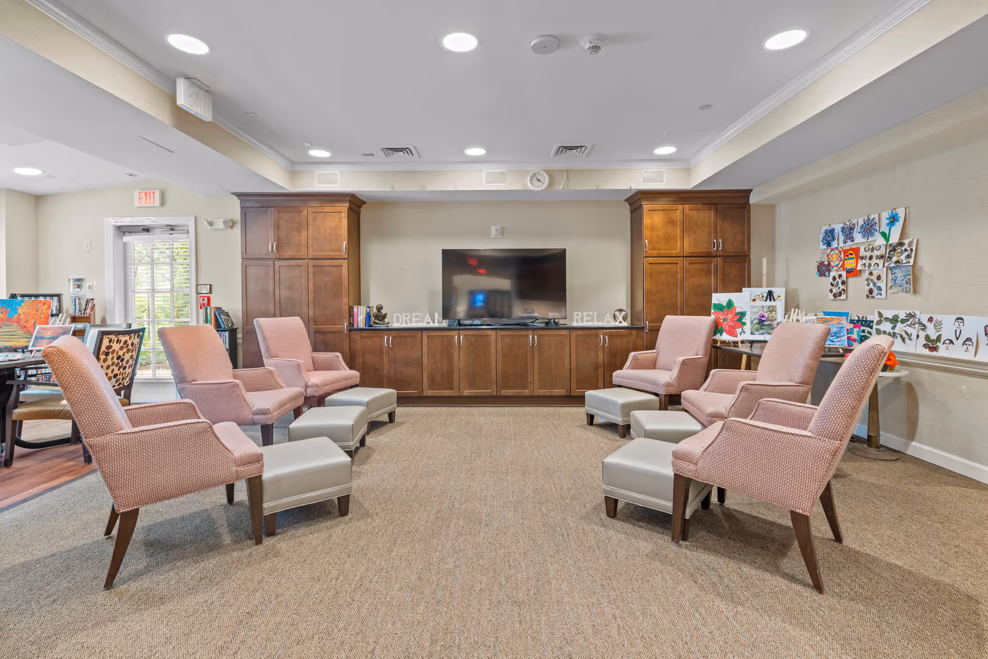 A senior living common room with pink armchairs and ottomans arranged facing a large TV set in wooden cabinets, with artwork on the walls.