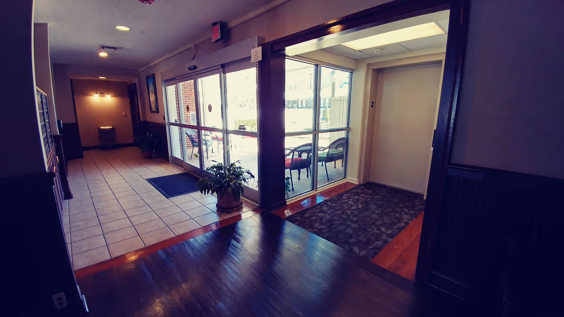 Interior view of an entrance area in a senior living facility with tiled flooring leading to glass double doors opening to an outdoor patio with chairs. To the right, there is an elevator with a patterned rug in front of it. The area is well-lit with ceiling lights and has some potted plants near the entrance.