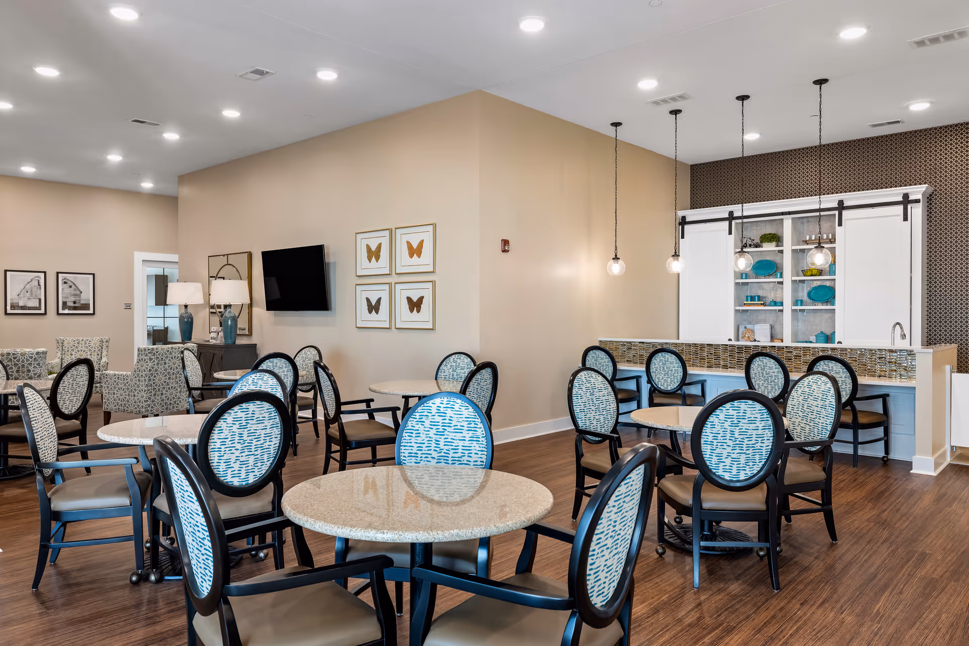 Bright communal dining room with round tables, patterned chairs, a serving counter with pendant lights, and a wall-mounted TV.