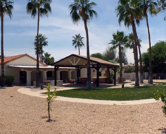 Outdoor area of a senior living facility with a covered patio featuring seating, surrounded by palm trees and a gravel landscape with a small patch of grass.