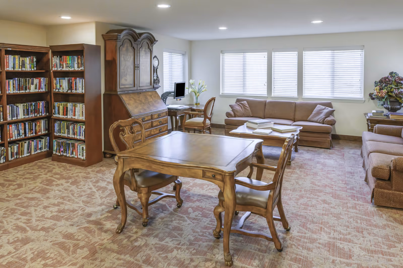 A cozy living room area with two brown sofas, a wooden coffee table with books on it, a wooden table with two chairs in the foreground, and bookshelves filled with books along the left wall. There is also a wooden cabinet with a clock and a computer desk with a chair near the windows with closed blinds.