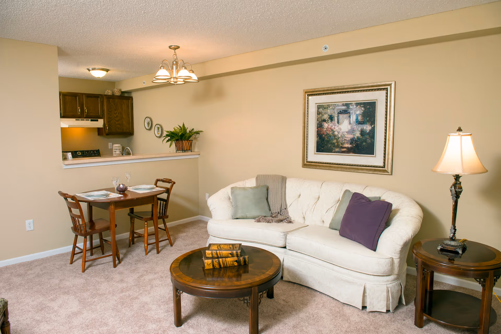 A cozy living room area with a cream-colored tufted sofa adorned with green and purple pillows, a round wooden coffee table with three decorative books, a wooden side table with a lamp, and a small dining table with two chairs set with plates and glasses. The background shows a partial view of a kitchen with dark wooden cabinets and a light fixture hanging from the ceiling. The walls are painted beige and there is a framed painting above the sofa.