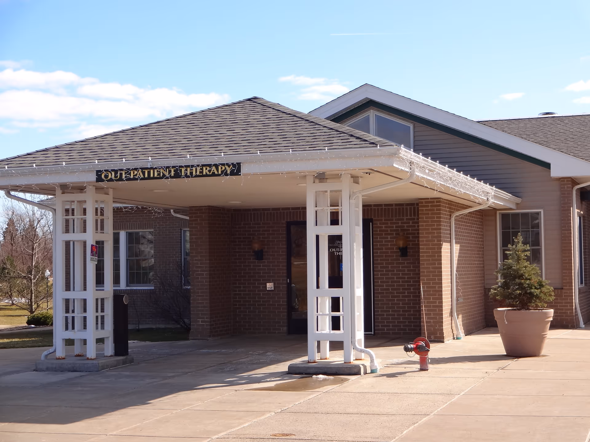 Covered front entrance and drive-up canopy of a care facility with a sign reading "Outpatient Therapy", a potted plant, and a fire hydrant connection.