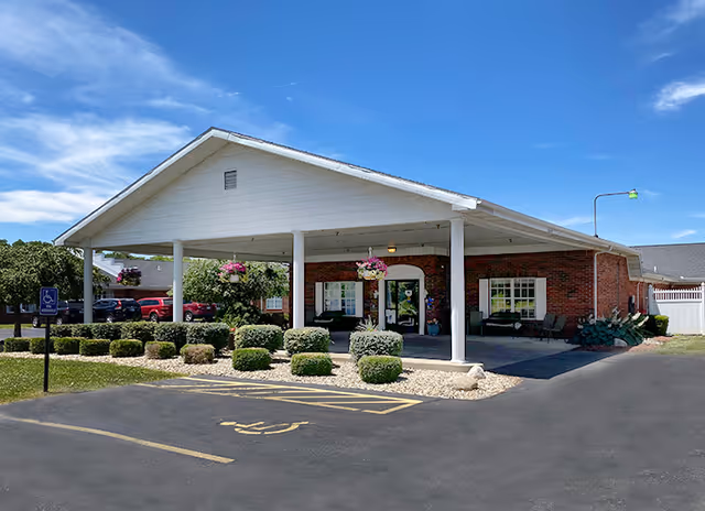 Front exterior view of a single-story brick building with a large covered entrance supported by white columns. There are hanging flower baskets under the covered area, neatly trimmed bushes in front, and a handicapped parking space visible in the foreground. The sky is clear and blue.