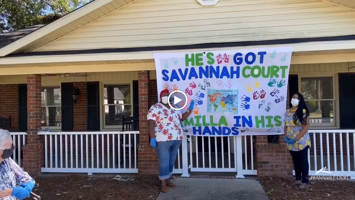 Two women wearing face masks and gloves stand outside a brick building with white railings, holding a large colorful banner that reads 'He's got Savannah Court Camilla in his hands' with handprints and a map of the world in the center. Another person wearing a mask and gloves is seated to the left.