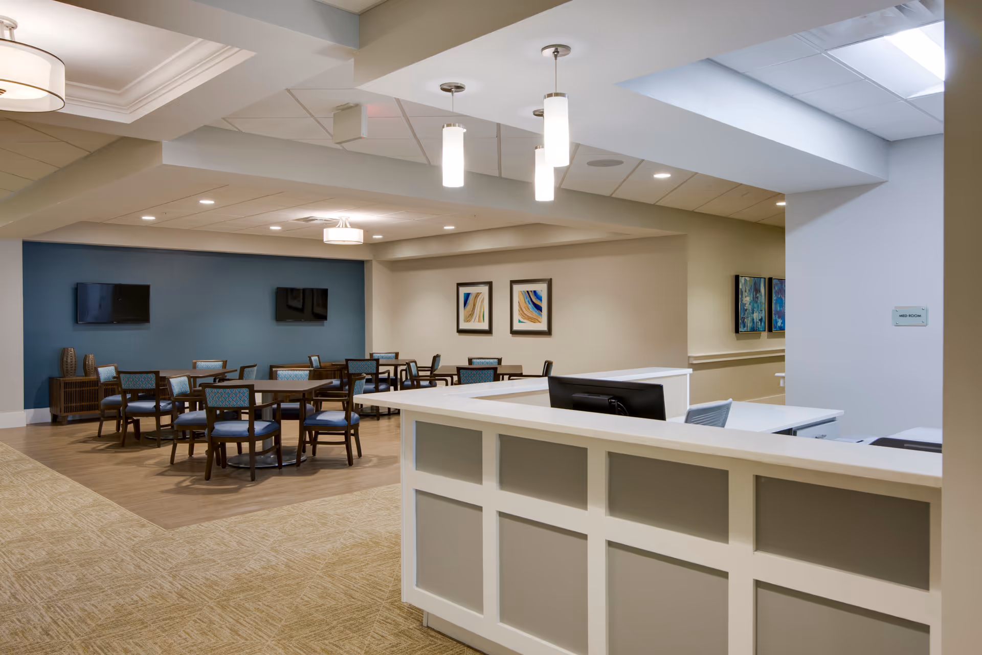 Reception desk in the foreground with a dining/lounge area of tables and chairs, wall-mounted TVs and artwork in a senior living facility.