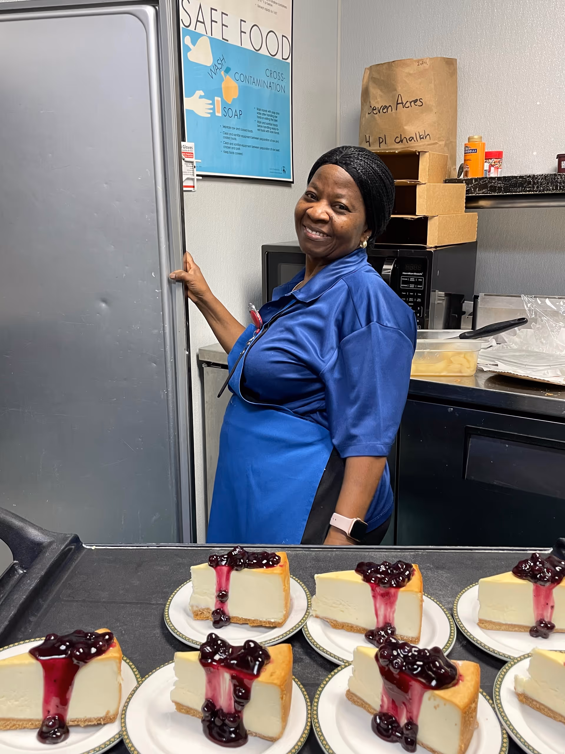 A smiling kitchen worker in a blue uniform stands behind a counter with several plates of cheesecake topped with berry sauce.