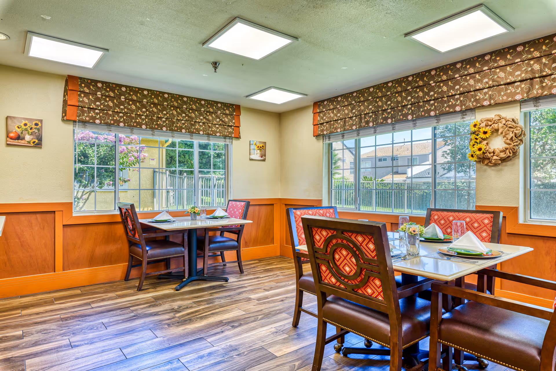 Bright dining room with two set tables and patterned chairs beside large windows overlooking greenery.