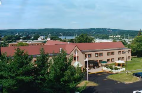 A beige multi-story senior living building with a red roof, trees, and a parking lot.