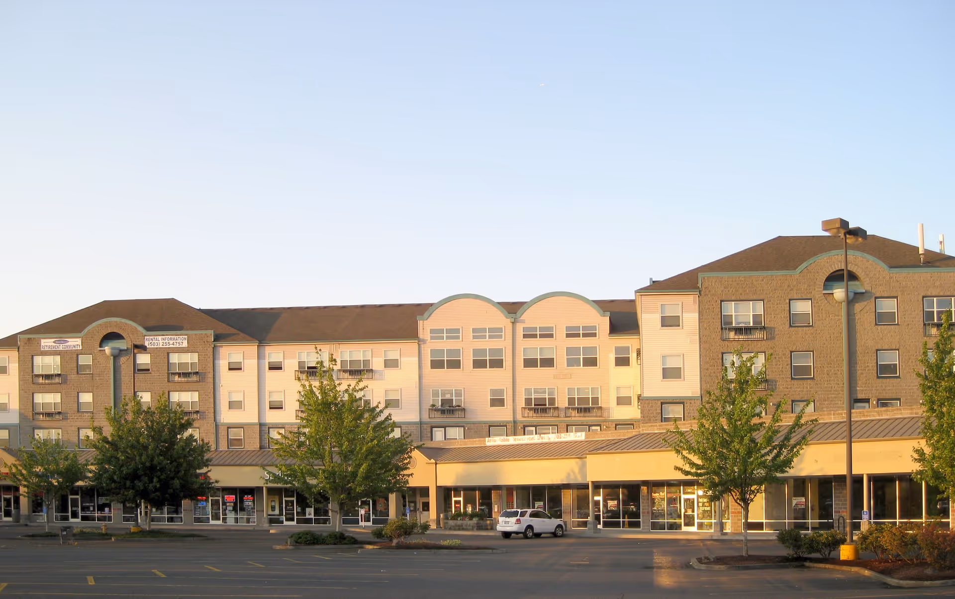 Exterior view of a multi-story building with retail shops on the ground floor and residential apartments above, surrounded by trees and a parking lot in front under a clear sky.