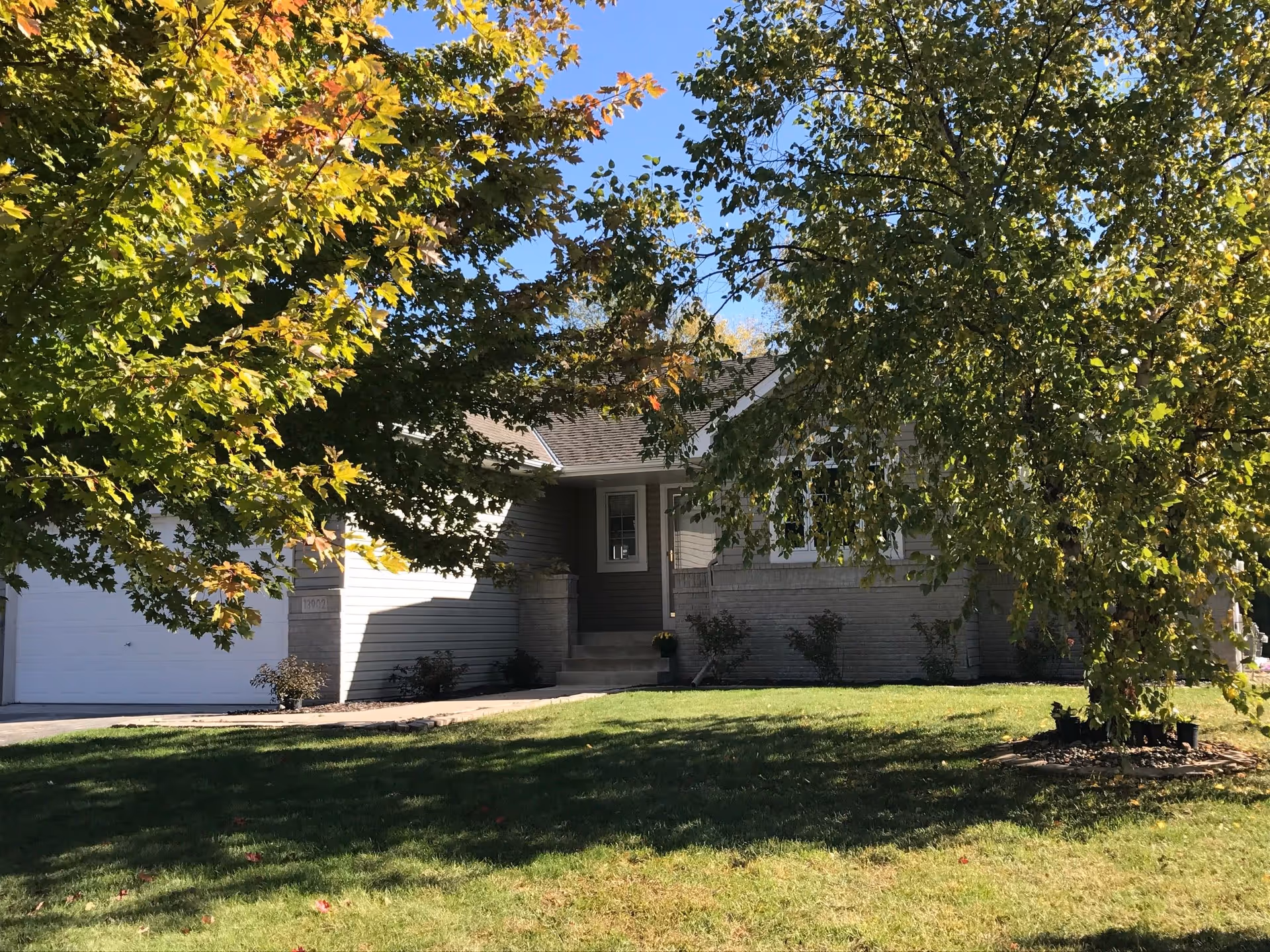 A single-story residential house partially obscured by green leafy trees in the front yard. The house has light-colored siding, a small set of steps leading to the front door, and a garage on the left side. The lawn is well-maintained with some shadows from the trees.